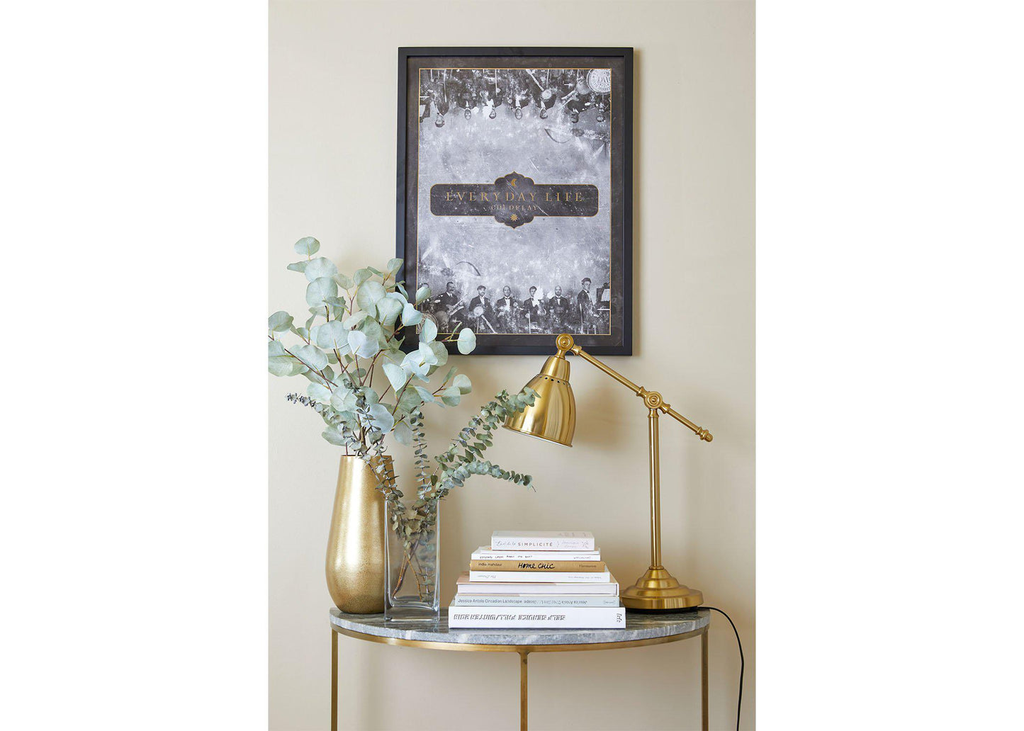 4: Everyday Life Lithograph on a black wooden frame, hung on a beige wall, above a small table shaped like a crescent moon, with a marble top and golden legs. The table has golden and clear vases with eucalyptus, books and a golden desk lamp. The artwork’s emphasis on golden themes embellish this modern space