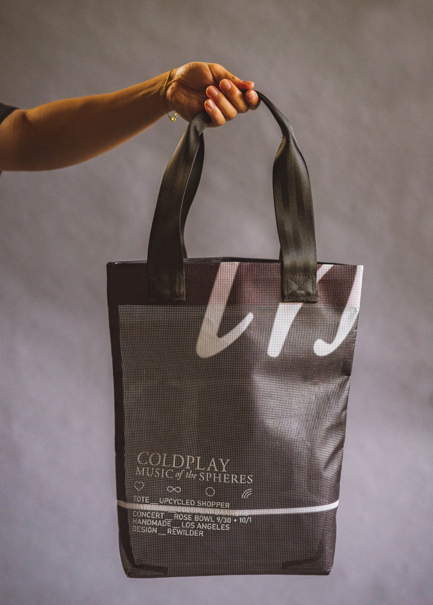 Black dark space tote bag. In white, COLDPLAY MUSIC OF THE SPHERES. A heart, an infinity sign and Chris’s rainbow doodle. Tote Upcycled Shopper. Material: Coldplay Banners. Concert: Rose Bowl. 9/30 & 10/1. Handmade. Los Angeles. Design: Rewilder. Some traces of writing are seen, but not readable