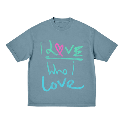 Front: The shirt is a soft green blue. The words I love Who I love are painted in a free, expressive brushstrokes fashion that suggests emotion and spontaneity. In the first I love, I&L are in light green. The O  is a pink fuchsia heart, drawn by hand. The rest of the first love word is light green. Who I love is light blue. The letters are creatively decorated, in an elegant style.