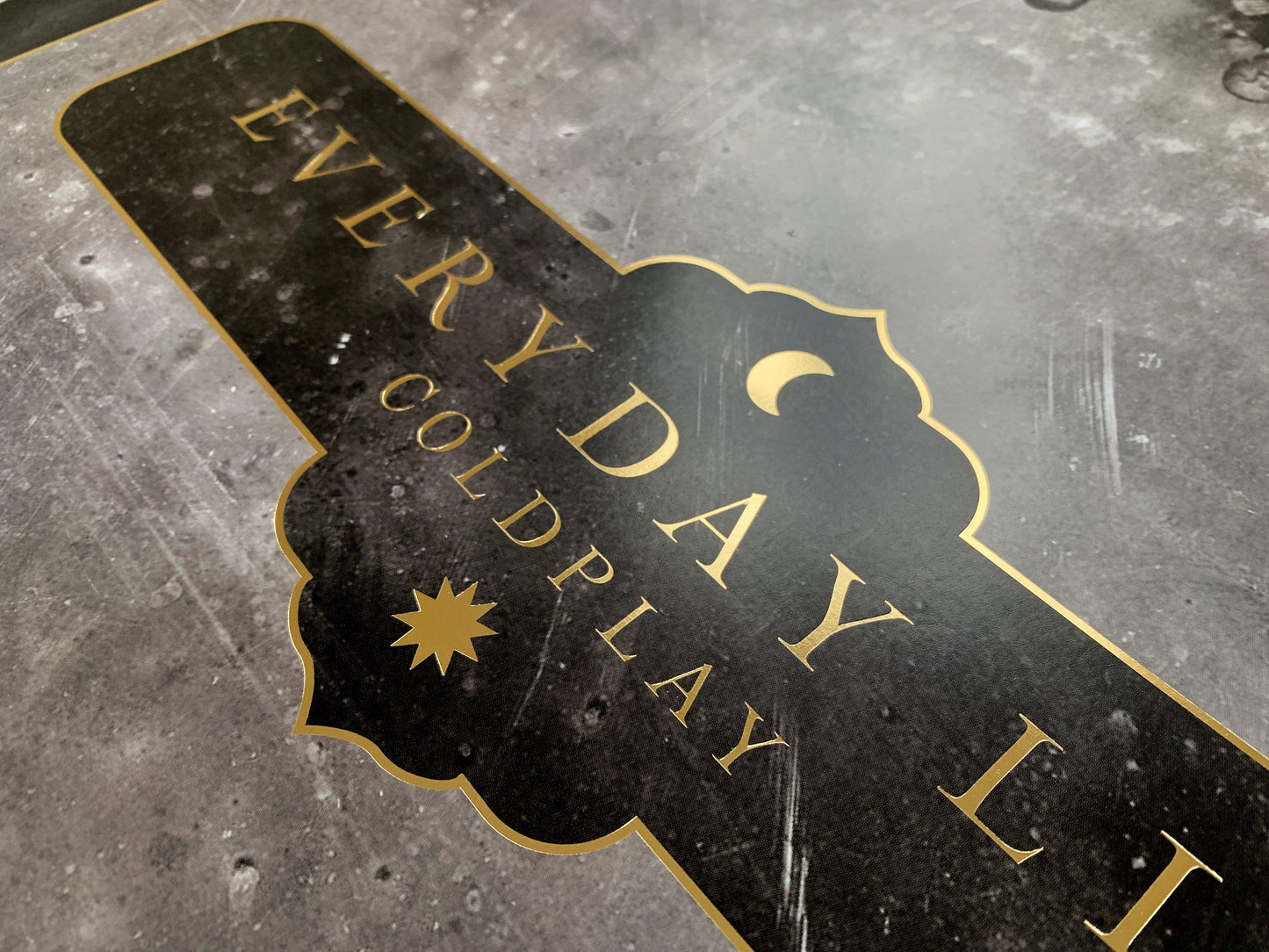 2: Lithograph text, close-up: Everyday Life and Coldplay printed in golden, with a crescent moon and a sun at the center. Both celestial bodies are golden. The text border is golden, ornate with curved edges, reminiscent of the peaks found in the arcs of mosques, or medieval shields. The Background of the poster is black