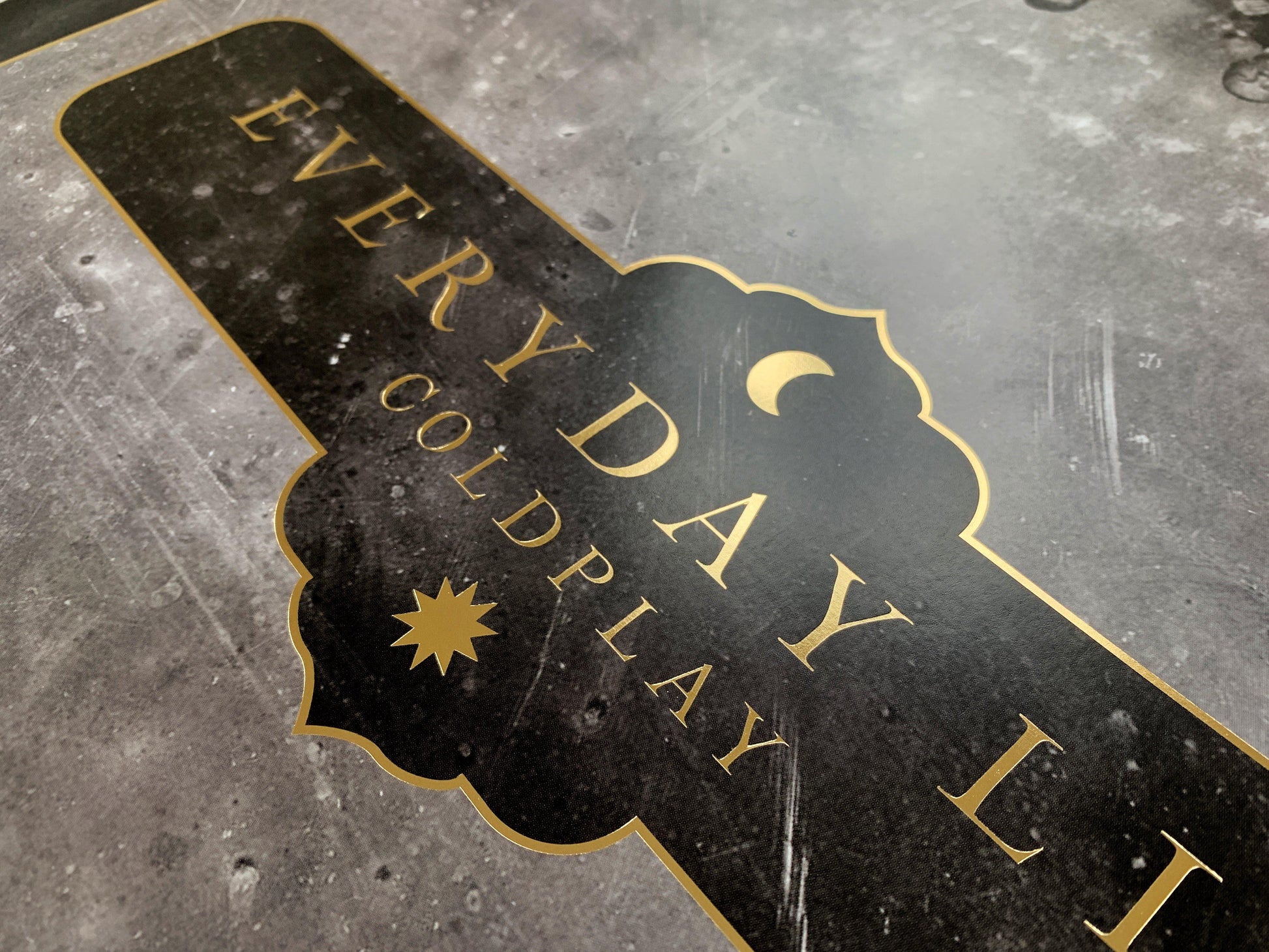 2: Lithograph text, close-up: Everyday Life and Coldplay printed in golden, with a crescent moon and a sun at the center. Both celestial bodies are golden. The text border is golden, ornate with curved edges, reminiscent of the peaks found in the arcs of mosques, or medieval shields. The Background of the poster is black