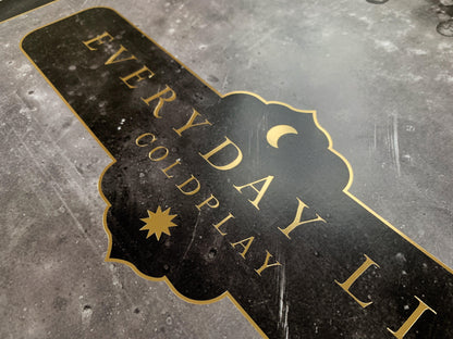 2: Lithograph text, close-up: Everyday Life and Coldplay printed in golden, with a crescent moon and a sun at the center. Both celestial bodies are golden. The text border is golden, ornate with curved edges, reminiscent of the peaks found in the arcs of mosques, or medieval shields. The Background of the poster is black