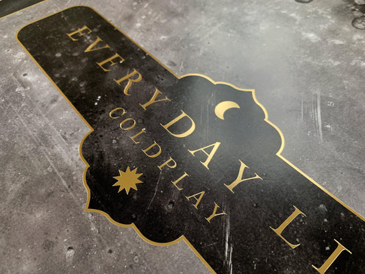 2: Lithograph text, close-up: Everyday Life and Coldplay printed in golden, with a crescent moon and a sun at the center. Both celestial bodies are golden. The text border is golden, ornate with curved edges, reminiscent of the peaks found in the arcs of mosques, or medieval shields. The Background of the poster is black