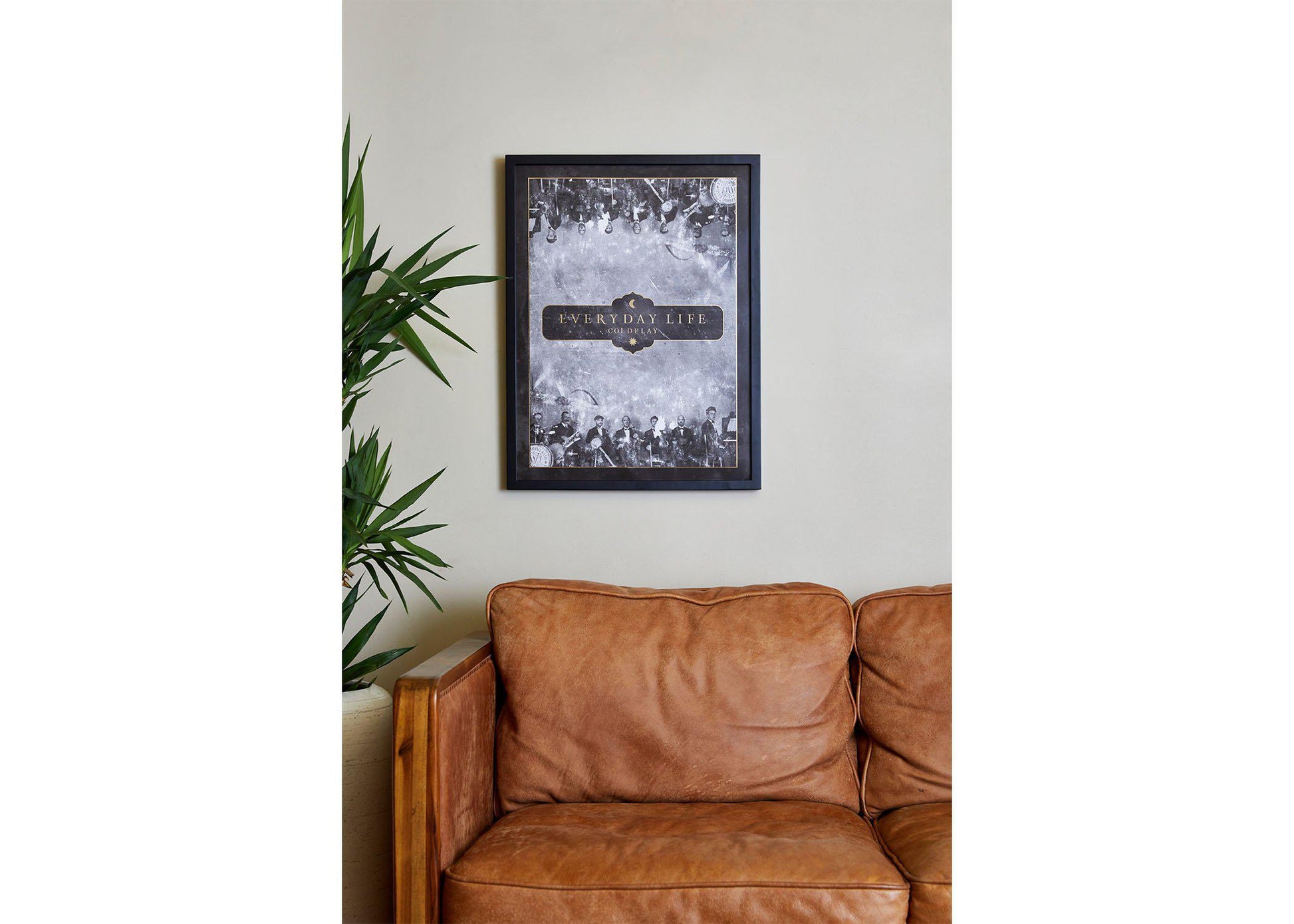 3: Everyday Life Lithograph in a cozy living room, above a vintage, caramel brown  leather sofa. To its left there’s a tall leafy plant in a pot, adding a touch of nature. The wall is painted beige. The poster has a black wooden frame. Black and white against the plant’s green make an elegant contrast
