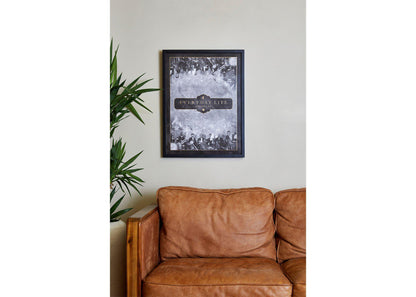 3: Everyday Life Lithograph in a cozy living room, above a vintage, caramel brown  leather sofa. To its left there’s a tall leafy plant in a pot, adding a touch of nature. The wall is painted beige. The poster has a black wooden frame. Black and white against the plant’s green make an elegant contrast