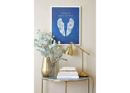 Image 4:
Ghost Stories lithograph on a white frame, hung on a beige wall, above a small table shaped like a crescent moon, with a marble top and golden legs. The table has golden and clear vases with eucalyptus, books and a golden desk lamp. The white angel wings add a touch of calm and serenity, complementing the room’s elegant decor