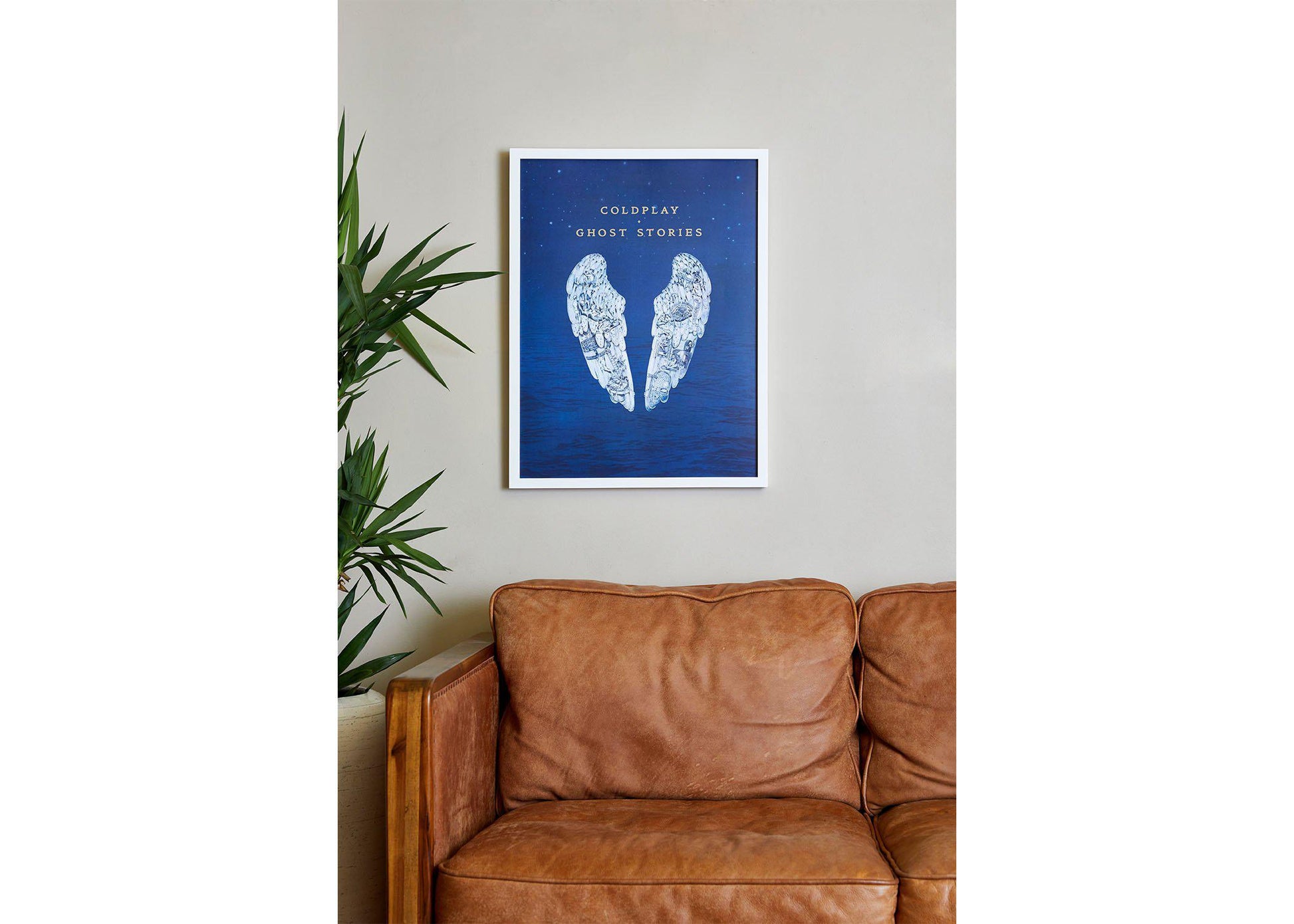Image 3:
Ghost Stories lithograph in a cozy living room, above a wooden, vintage, caramel brown leather sofa. To its left there’s a tall leafy plant in a pot, adding a touch of nature. The wall is beige. The poster is on a white frame. The artwork’s deep blue adds a splash of color to the space
