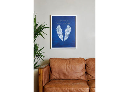 Image 3:
Ghost Stories lithograph in a cozy living room, above a wooden, vintage, caramel brown leather sofa. To its left there’s a tall leafy plant in a pot, adding a touch of nature. The wall is beige. The poster is on a white frame. The artwork’s deep blue adds a splash of color to the space