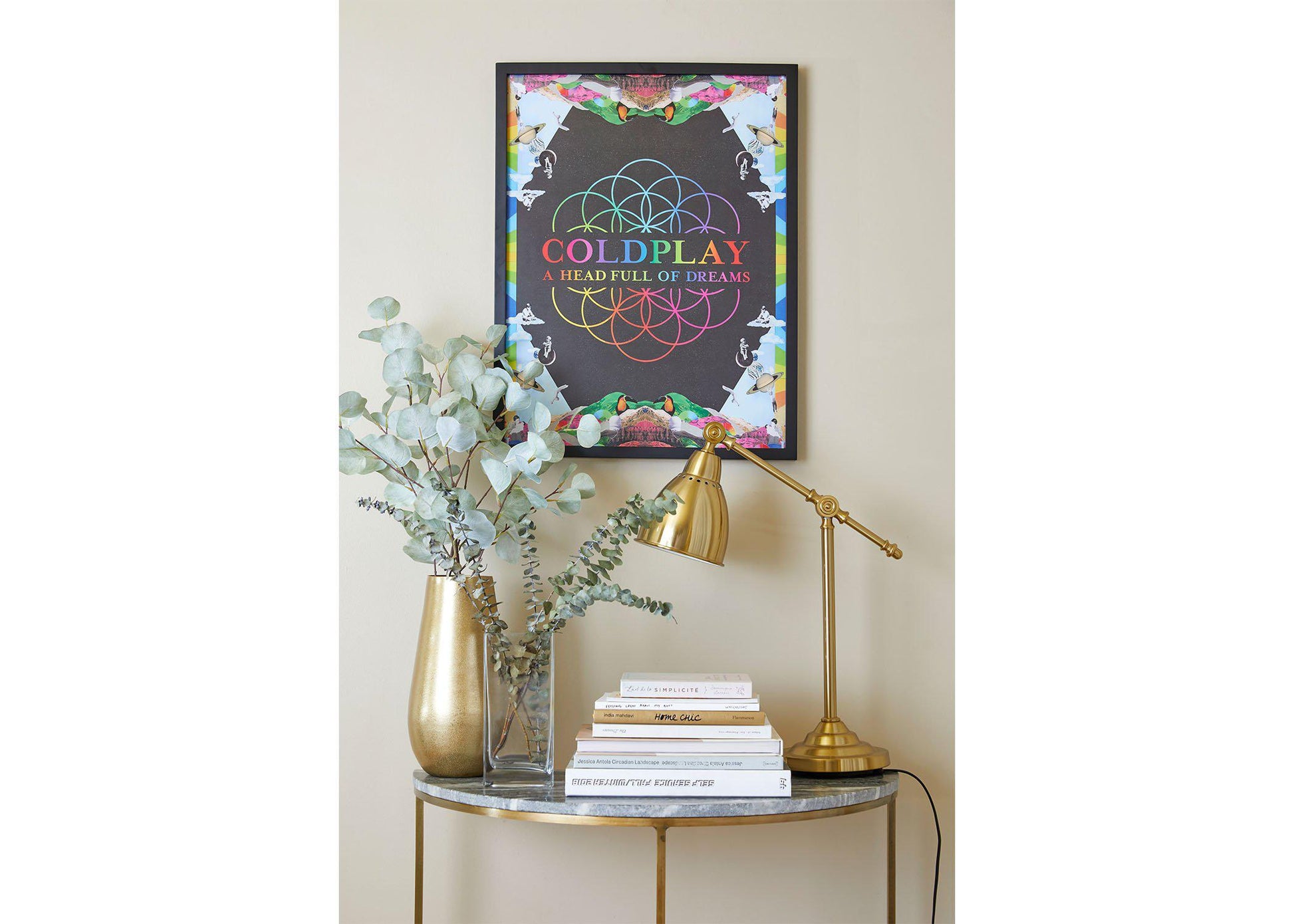 Image 5:
A Head Full Of Dreams Lithograph, hung above a small table shaped like a crescent moon, with a marble top and golden legs. The table has golden and clear vases with eucalyptus, books and a golden desk lamp. The poster is on a black wooden frame. The colorful artwork contrasts with the beige wall