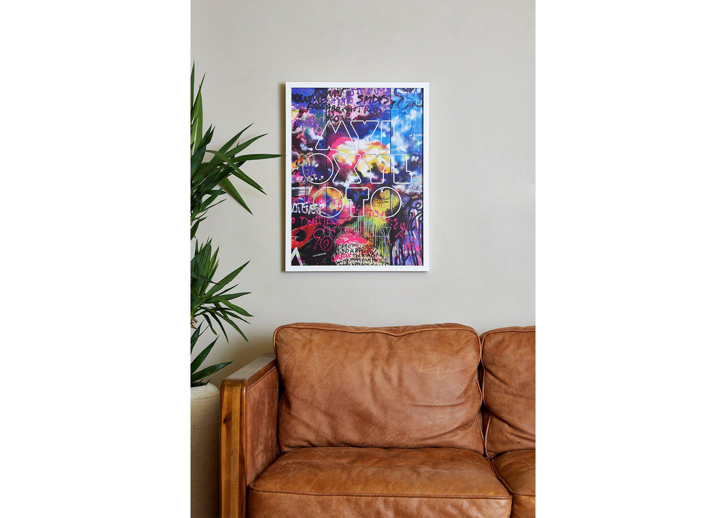 Image 3:
Mylo Xyloto lithograph on a white frame in a cozy living room, hung above a vintage caramel brown leather sofa. There’s a tall leafy plant in a pot, adding a touch of nature. The blues, reds, yellows and purples stand out against the beige wall. Its graffiti gives the room a bold, contemporary feel
