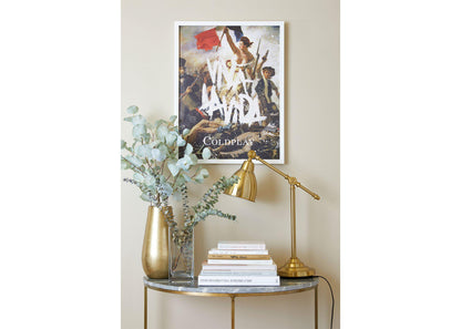 Image 4:
Viva La Vida Lithograph on a white frame, hung on a beige wall, above a small table shaped like a crescent moon, with a marble top and golden legs. The table has golden and clear vases with eucalyptus, books and a golden desk lamp. The painting’s references to art and music give the room a dynamic, cultured feel