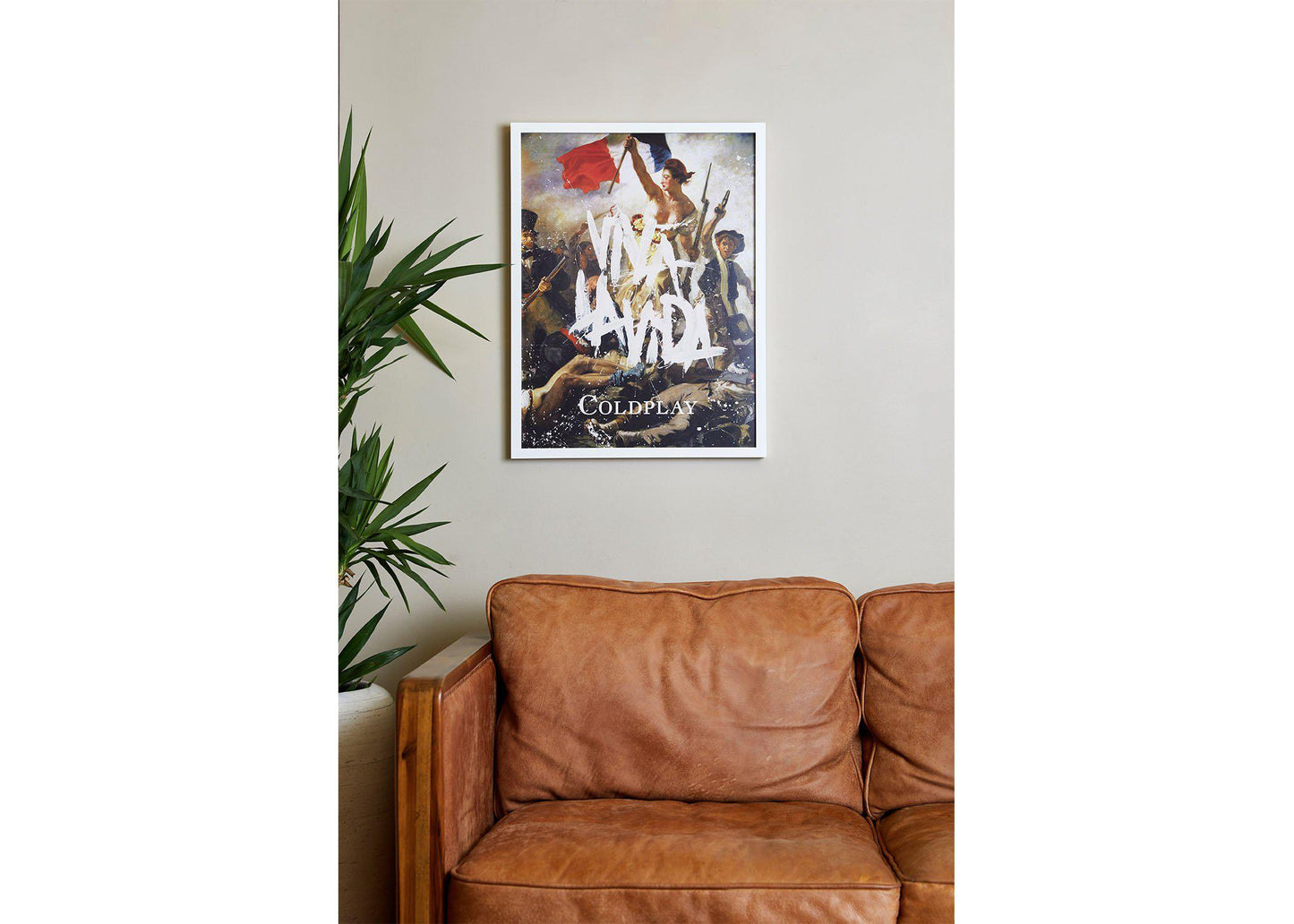 Image 3:
Viva La Vida Lithograph in a cozy living room, hung above a wooden, vintage caramel brown leather sofa. To its left, there’s a tall leafy plant in a pot, adding a touch of nature. The wall is beige. The poster is on a white frame. The text’s raw, rebellious brushstrokes evoke creativity and energy