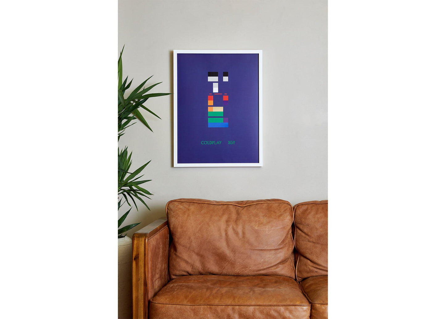 Image 3:
X&Y Lithograph on a white frame, in a cozy living room, hung above a wooden, vintage, caramel brown leather sofa. To its left, there’s a tall leafy plant in a pot. The wall is beige. The colorful blocks make the neutral room dynamic and lively. The green on them subtly relates to the green on the plant